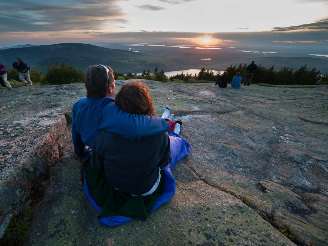 While seated on a blanket on a patch of mountaintop granite, a man and woman clasp their arms around