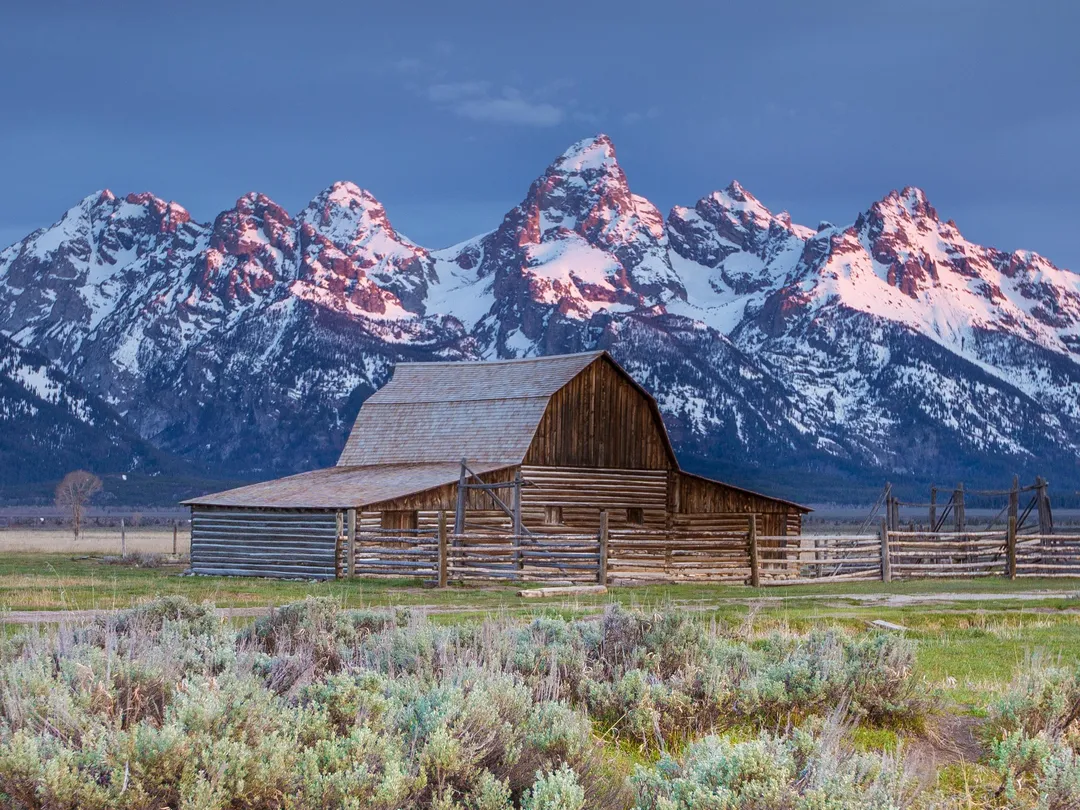 A wooden barn with snowy peaks in the background and green vegetation in the foreground