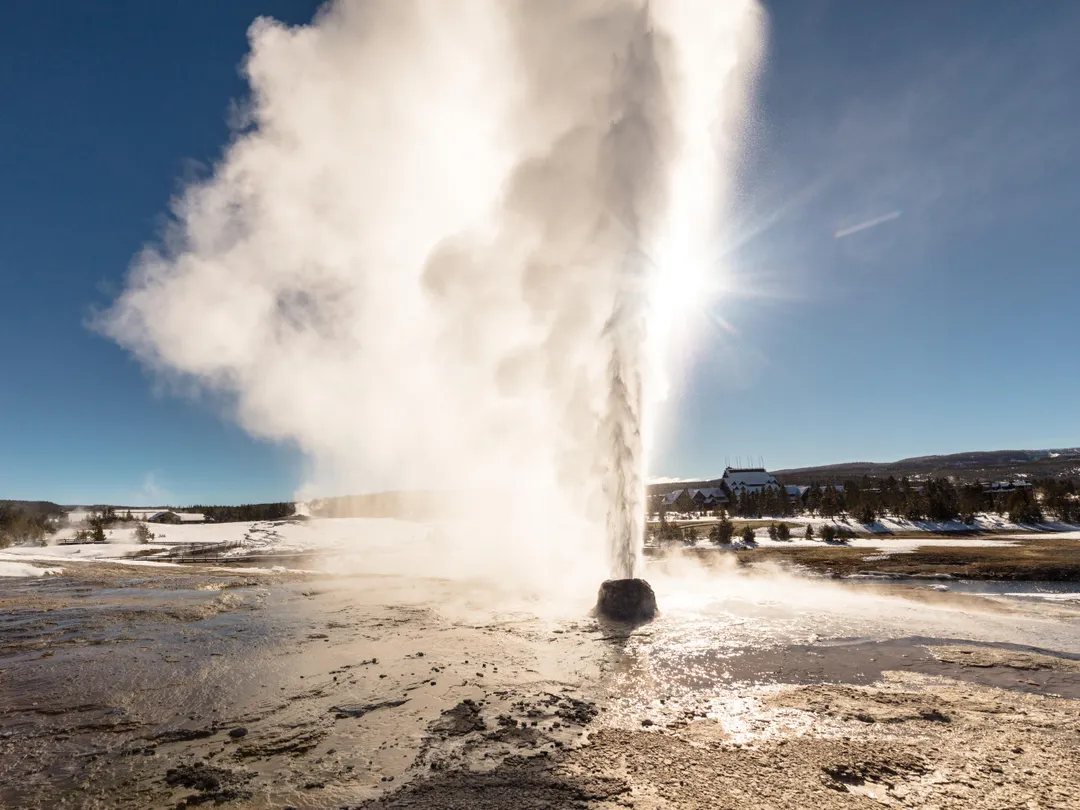 A geyser erupts steam in front of the bright sun.